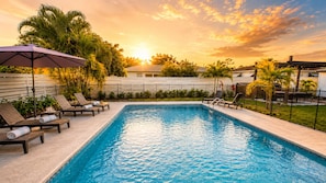 Ein rechteckiger Swimmingpool mit Liegestühlen und Sonnenschirmen, umgeben von einem Zaun und Palmen.