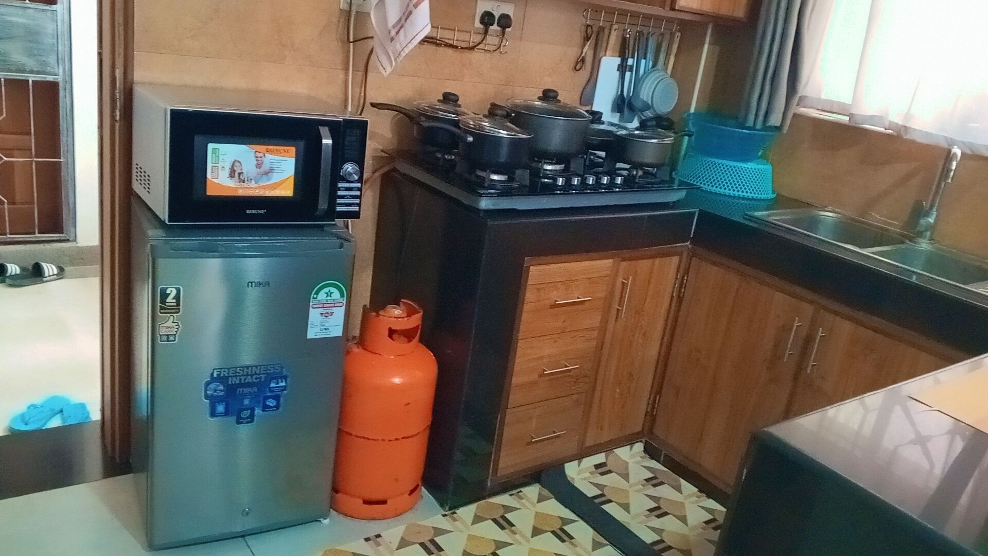 A compact kitchen with a refrigerator, stove, gas cylinder, and a microwave.