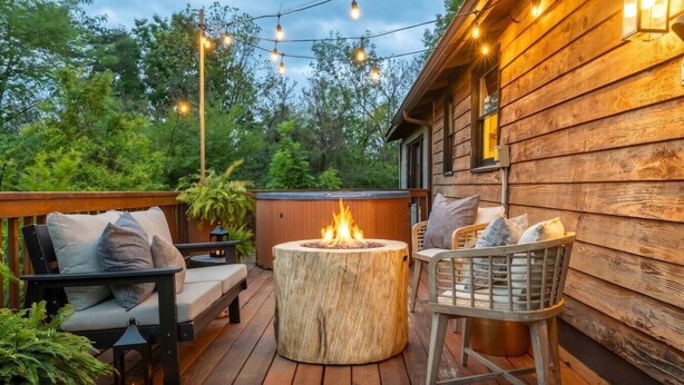 Una terraza de madera con mobiliario exterior, una hoguera, luces colgantes y vistas a árboles.