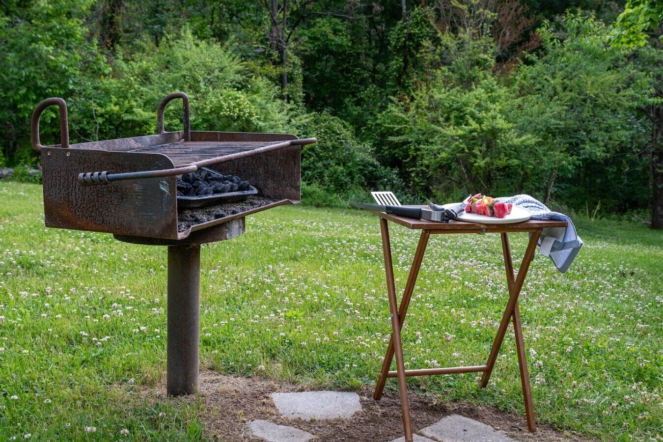 A barbecue grill with charcoal, a portable table with a plate of food, and a grill brush.