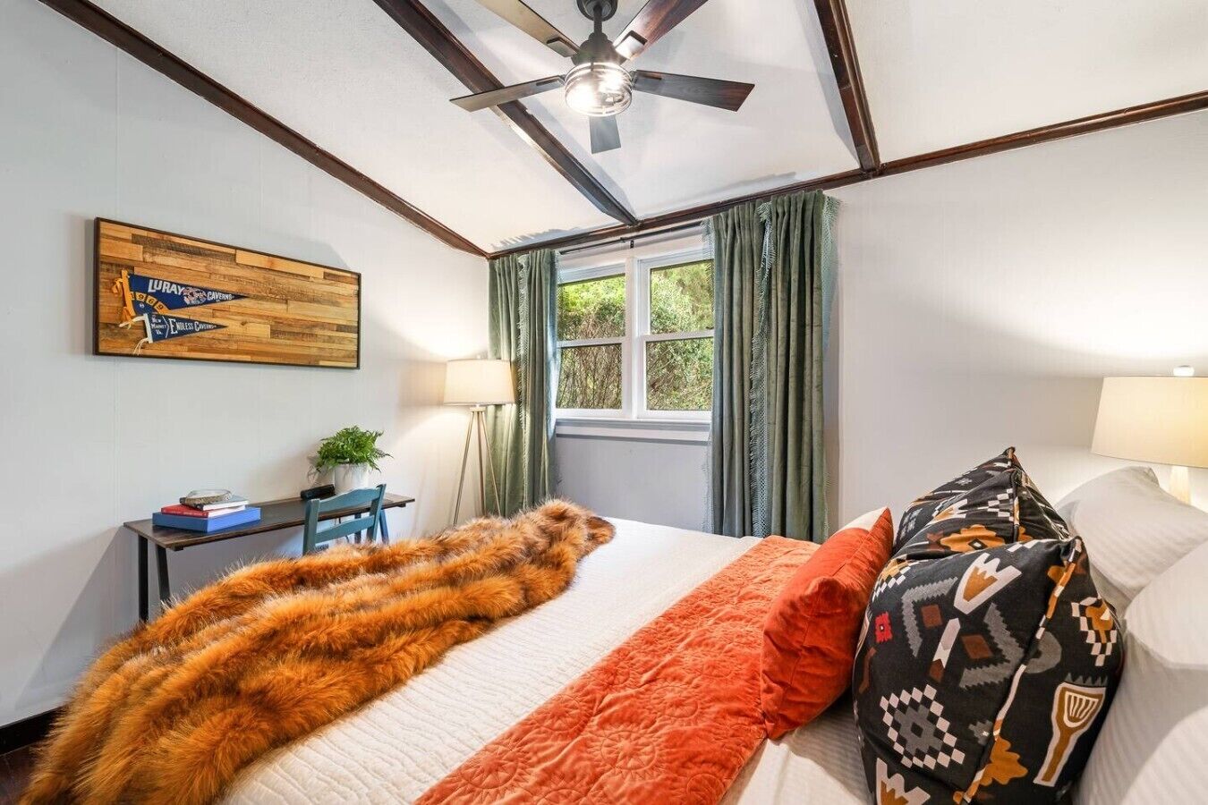 Une chambre d’hôtel comprenant un lit, un ventilateur de plafond, une fenêtre avec des rideaux, un bureau avec une lampe et une œuvre d’art accrochée au mur.