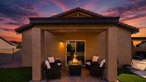 A covered patio with two chairs and a table, a fire feature, and a view of the sunset. - Luxury Private Pool & Spa Home | Resort Backyard | Sleeps 10 (Tucson)