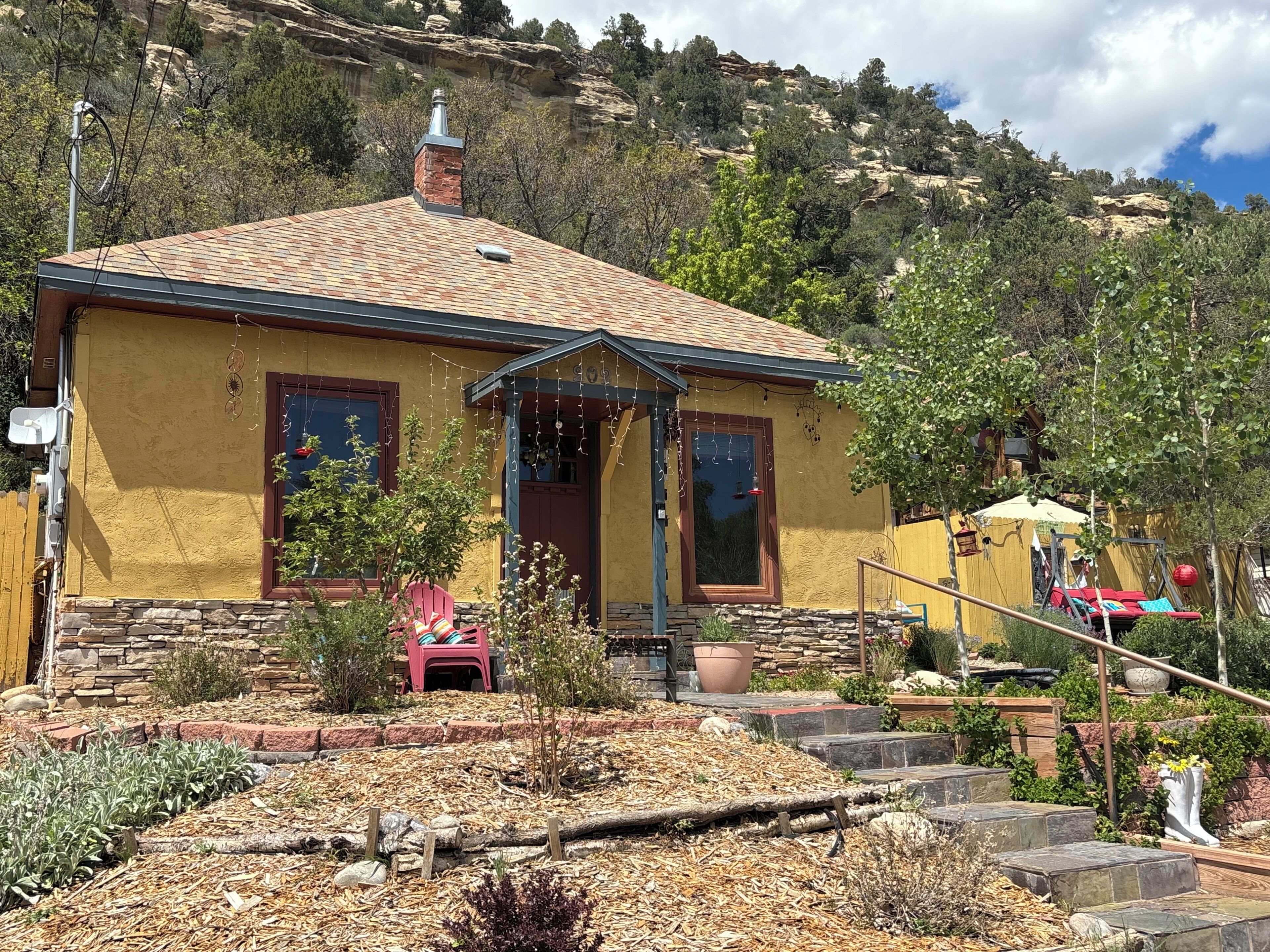 A yellow building with a brick chimney, a porch with a red chair, and a garden with mulch and plants.