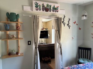 Quarto com cama, estante de madeira com objetos decorativos, janela com cortinas, TV fixada na parede e uma obra de arte com tema de cactos.