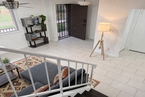 Ein Wohnzimmer mit grauem Sofa, einem Holztisch, einem gemusterten Teppich, einem Bücherregal, einer Tür mit einem Metallgitter und einer Stehlampe.
