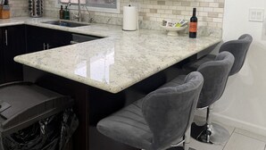 Una cocina con armarios oscuros, encimera de mármol, un lavamanos, una ventana y taburetes de bar.