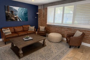 Ein Wohnzimmer mit braunem Ledersofa, einem Holztisch, einem runden Hocker und einer Ziegelwand.