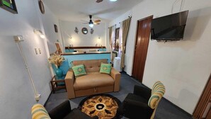 A living room with a sofa, two armchairs, a coffee table, a ceiling fan, a TV, and a kitchen area in the background. - Mediterranea Hostel (Córdoba)