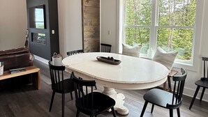Ein moderner Essbereich mit einem runden Tisch, schwarzen Stühlen, einer Pendelleuchte und einem großen Fenster mit Blick auf Bäume.