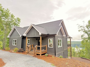 Ein zweistöckiges Haus mit Veranda, einem Schotterweg und Blick auf einen See und Wald.