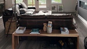 Ein geräumiges Wohnzimmer mit hoher Decke, großen Fenstern, einem gemütlichen Ecksofa, einem Holztisch und einem Fernseher.