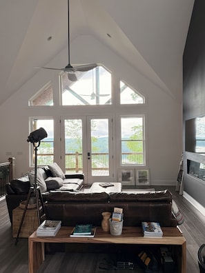 Ein geräumiges Wohnzimmer mit hoher Decke, großen Fenstern, einem gemütlichen Ecksofa, einem Holztisch und einem Fernseher.