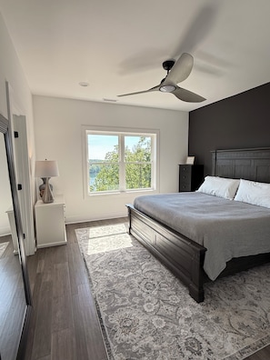 Ein Schlafzimmer mit einem Bett, Deckenventilator, Nachttisch, Lampe, Fenster mit Blick und einem gemusterten Teppich.