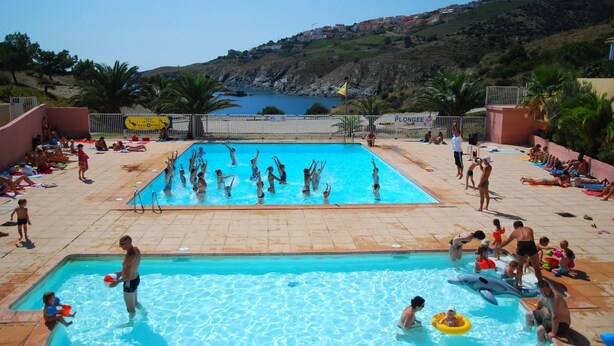 Ein Schwimmbecken mit darin badenden Personen, umgeben von einer Terrasse mit Stühlen und Tischen.