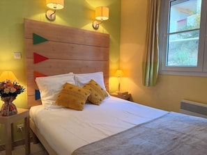 A bed with white bedding, two decorative pillows, a wooden headboard with directional arrows, two wall-mounted lamps, a window with curtains, and a vase with flowers on a bedside table. - Le Village des Aloes et des Bruyeres (Cerbère)