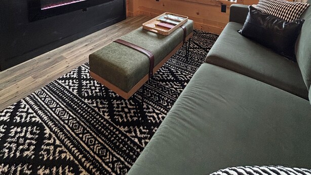 Interior acolhedor de cabana com uma TV grande fixada na parede, um sofá confortável, teto de madeira e uma janela com vista para o exterior.
