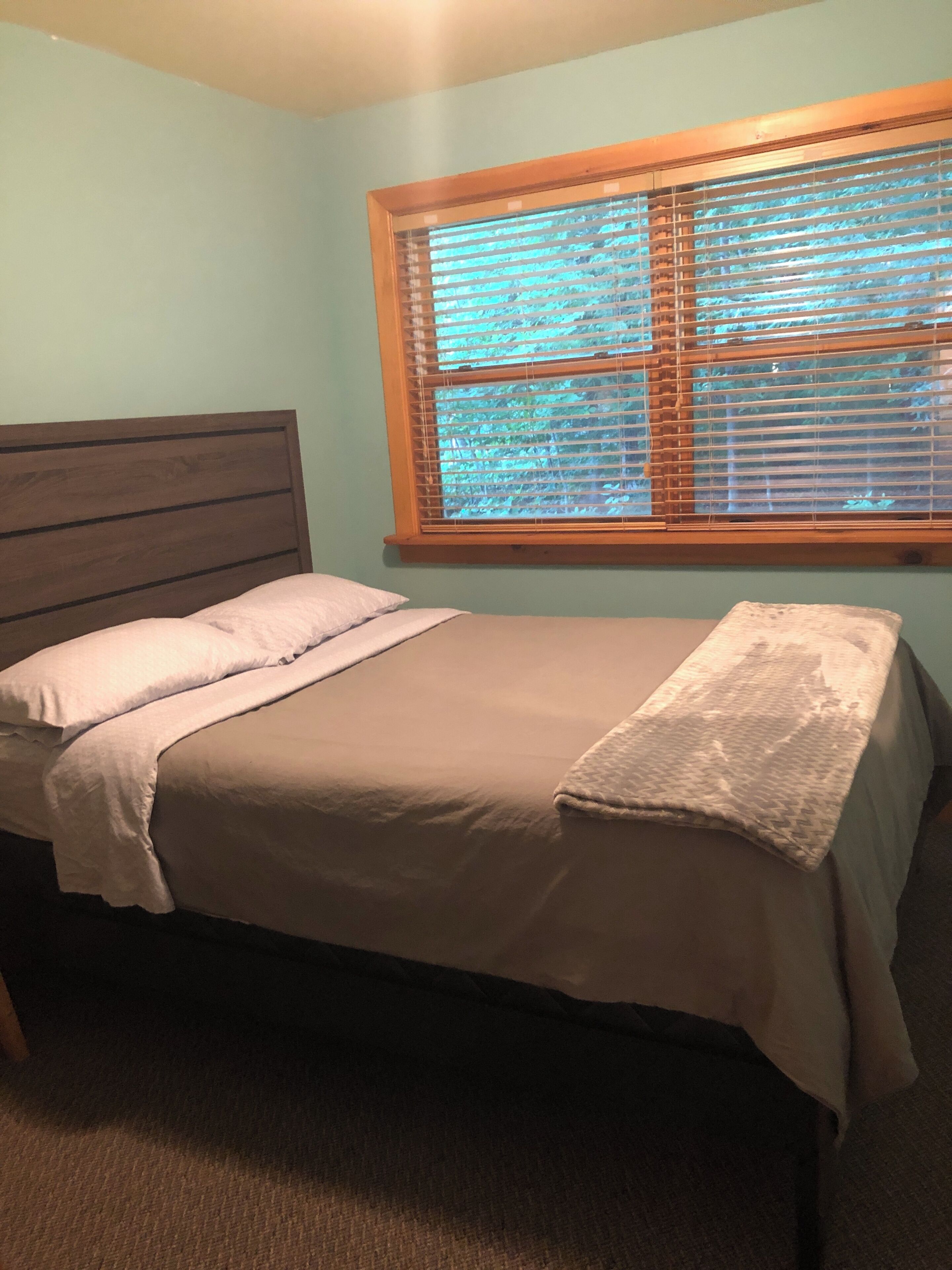 Une chambre avec une tête de lit en bois, un lit simple avec une couvre-lit et des oreillers de couleur beige, une fenêtre avec des stores et un sol recouvert d'une moquette.