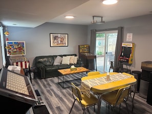 Una sala de estar con un sofá de cuero negro, una mesa de centro, una mesa amarilla con sillas, una máquina de pinball y un cuadro en la pared.