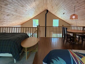A loft bedroom with a wooden ceiling, a bed, a chair, a dining table with chairs, and two windows.