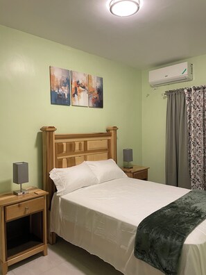Ein Schlafzimmer mit einem Bett, zwei Nachttischen, einer Klimaanlage und einem Fenster mit Vorhängen.