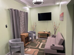 Ein Wohnzimmer mit einer Couch, zwei Sesseln, einem kleinen Tisch, einem Flachbildfernseher und Zimmerpflanzen.
