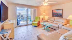 A living room with a sofa, a green chair, a table, a TV, and a balcony with a view. - Oceanview Obsession - 2Bdr Condo w/Pool & Hottub (New Smyrna Beach)