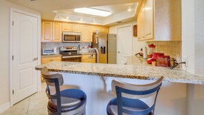 A kitchen with granite countertops, stainless steel appliances, wooden cabinets, and two bar stools. - Oceanview Obsession - 2Bdr Condo w/Pool & Hottub (New Smyrna Beach)