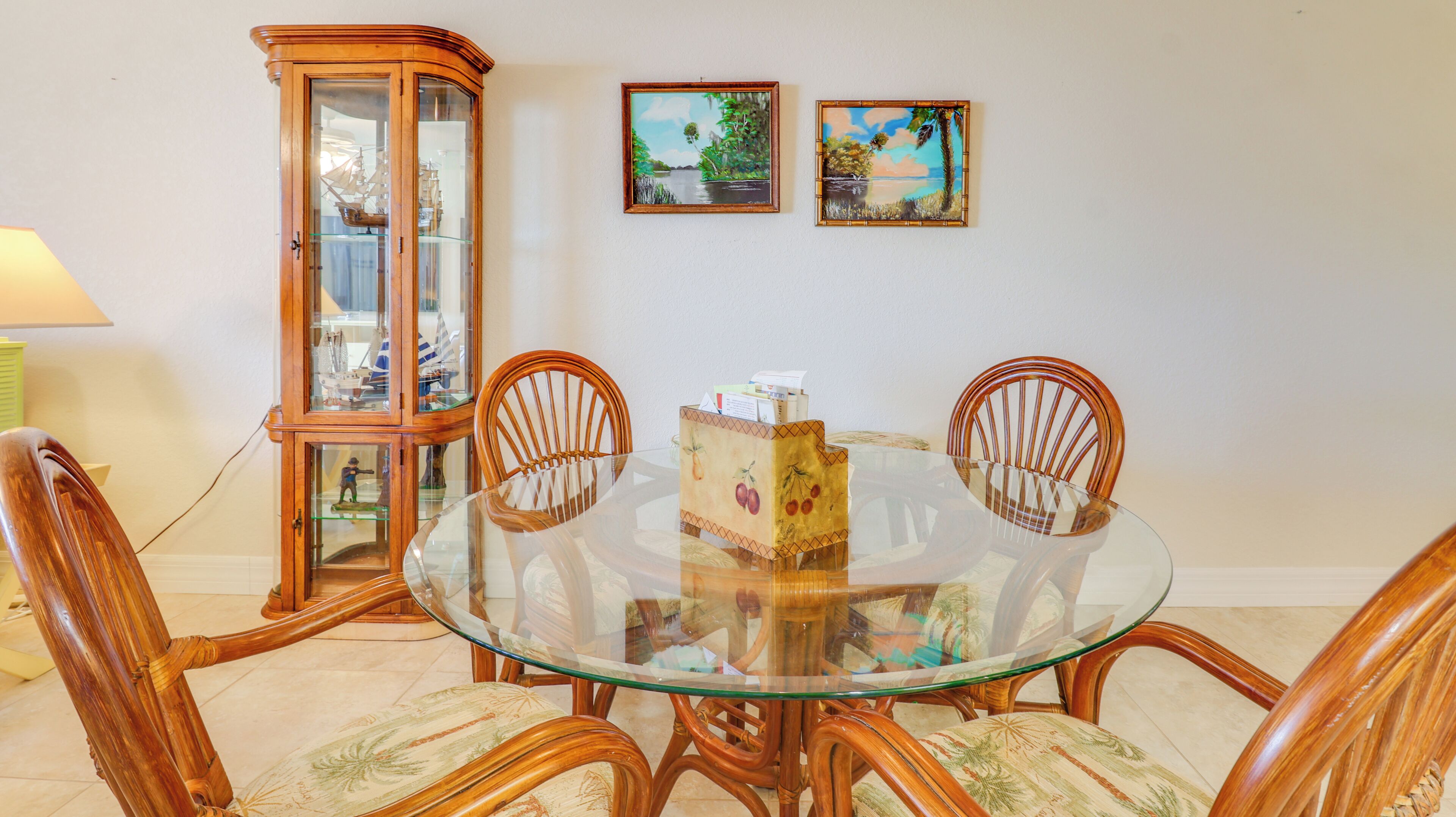 Un comedor con una mesa de vidrio rodeada de sillas de madera, un aparador de madera con puertas de cristal y dos cuadros de paisajes enmarcados en la pared.