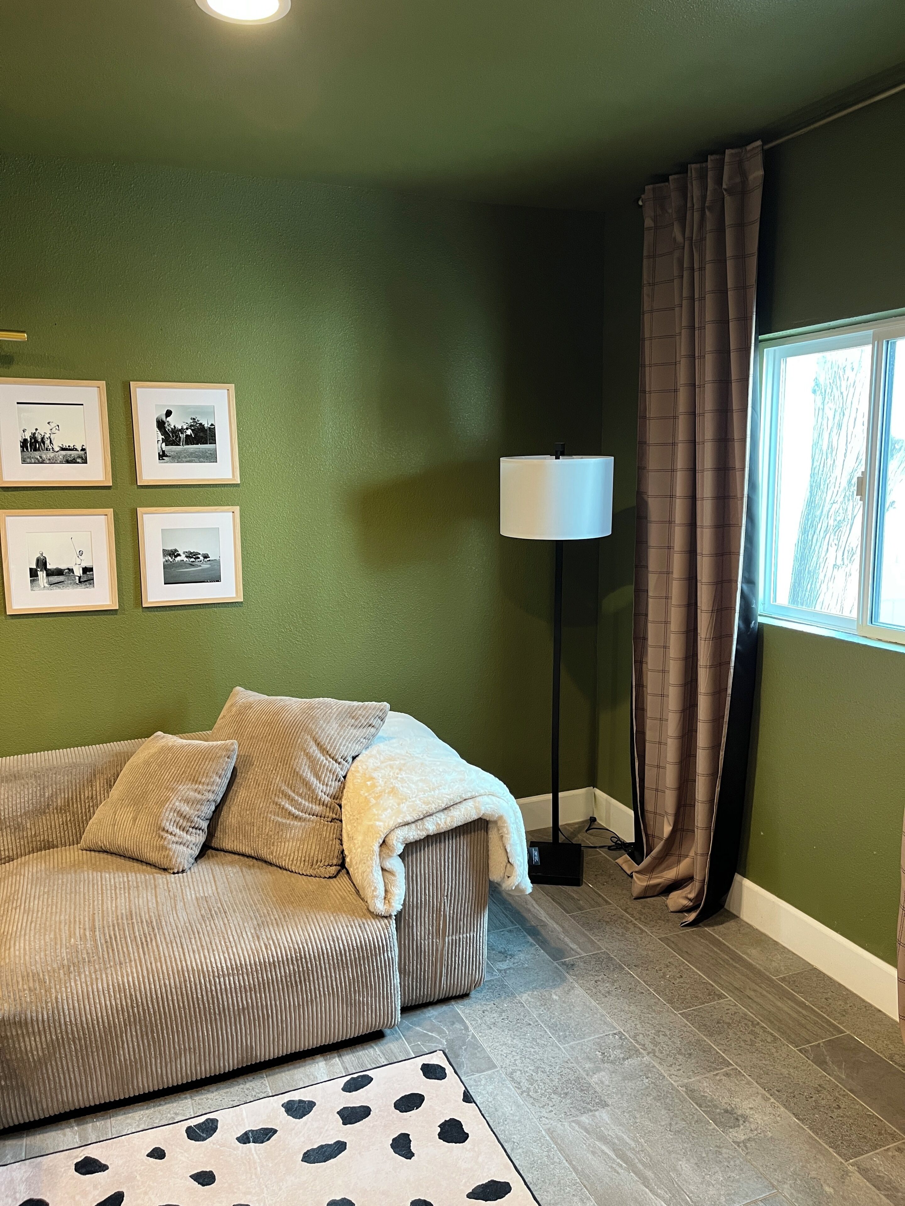A room with a green wall, a sofa, a floor lamp, a window with curtains, and framed pictures.