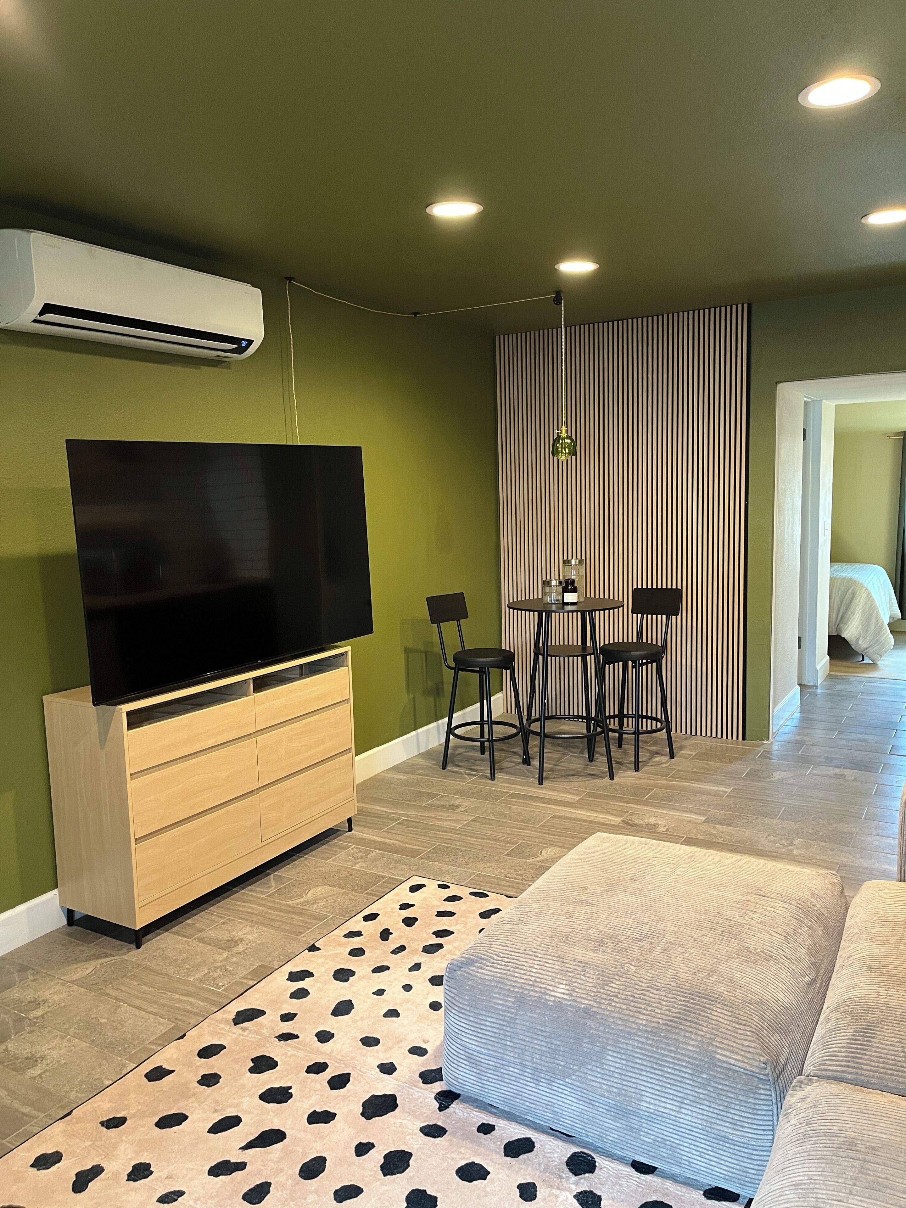 A living room with a flat-screen TV, wooden entertainment unit, a patterned rug, a sofa, a dining area with a table and chairs, and a striped partition.
