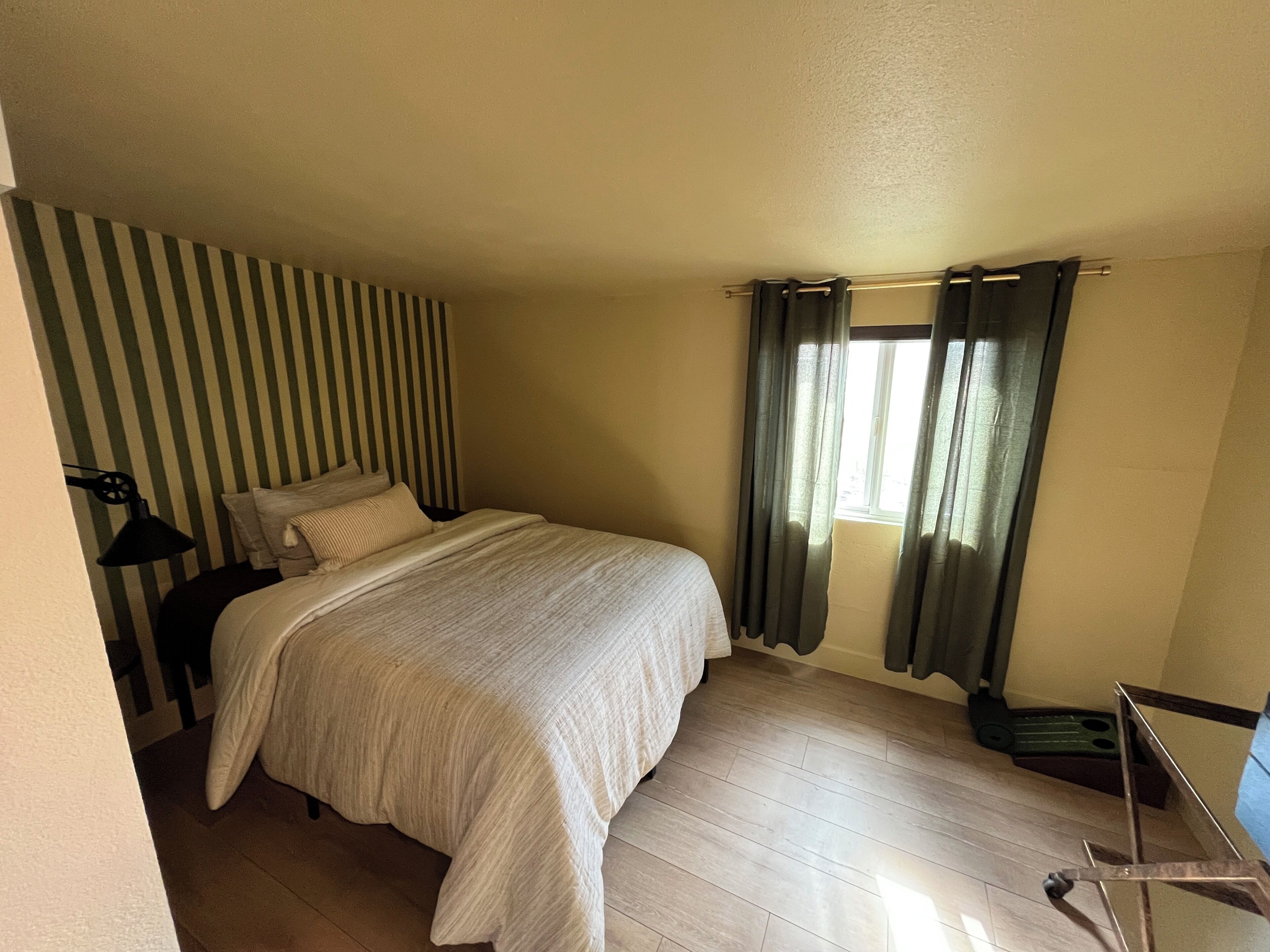 A bedroom with a bed, a nightstand, a lamp, a window with curtains, and a wooden floor.