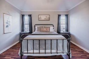Une chambre à coucher avec un lit en métal, une tête de lit, une table de chevet avec une lampe et une fenêtre avec des rideaux.