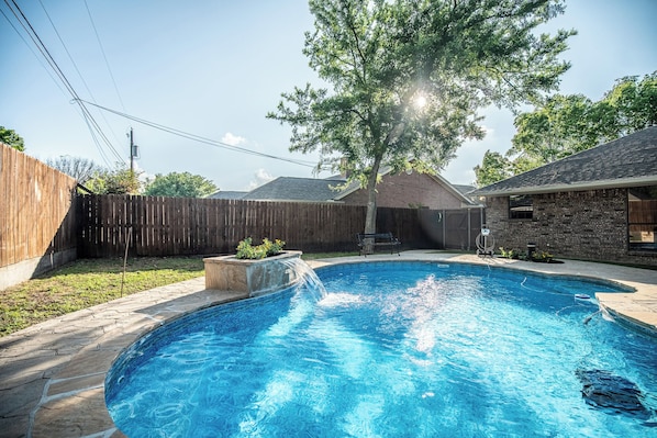 A backyard with a swimming pool, a wooden fence, a tree, and a stone patio.