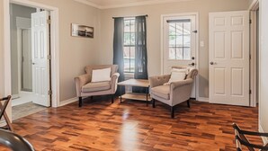 Ein Zimmer mit zwei Sesseln, einem kleinen Tisch, Holzdielen und einer Tür.