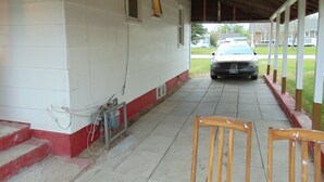 A covered porch with a car parked outside, a wooden chair, and a building with a red baseboard and white walls. - Just You Vacation Home - Main floor use (Gilbert Plains)