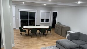 Ampla sala de estar com piso de madeira, sofá seccional cinza, mesa de jantar com cadeiras e área de cozinha com armários.