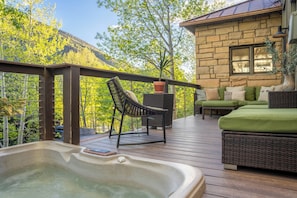 A wooden deck with a hot tub, wicker furniture, and a stone building with a window. - Luxury Remodeled 4BR Telluride Home | Hot Tub, Walk to Gondola & Main St | Sleeps 12 (Telluride)