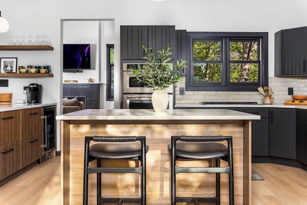 A modern kitchen with dark cabinetry, a large island with two stools, a TV mounted above the island, and a window with a view of trees. - Luxury Remodeled 4BR Telluride Home | Hot Tub, Walk to Gondola & Main St | Sleeps 12 (Telluride)