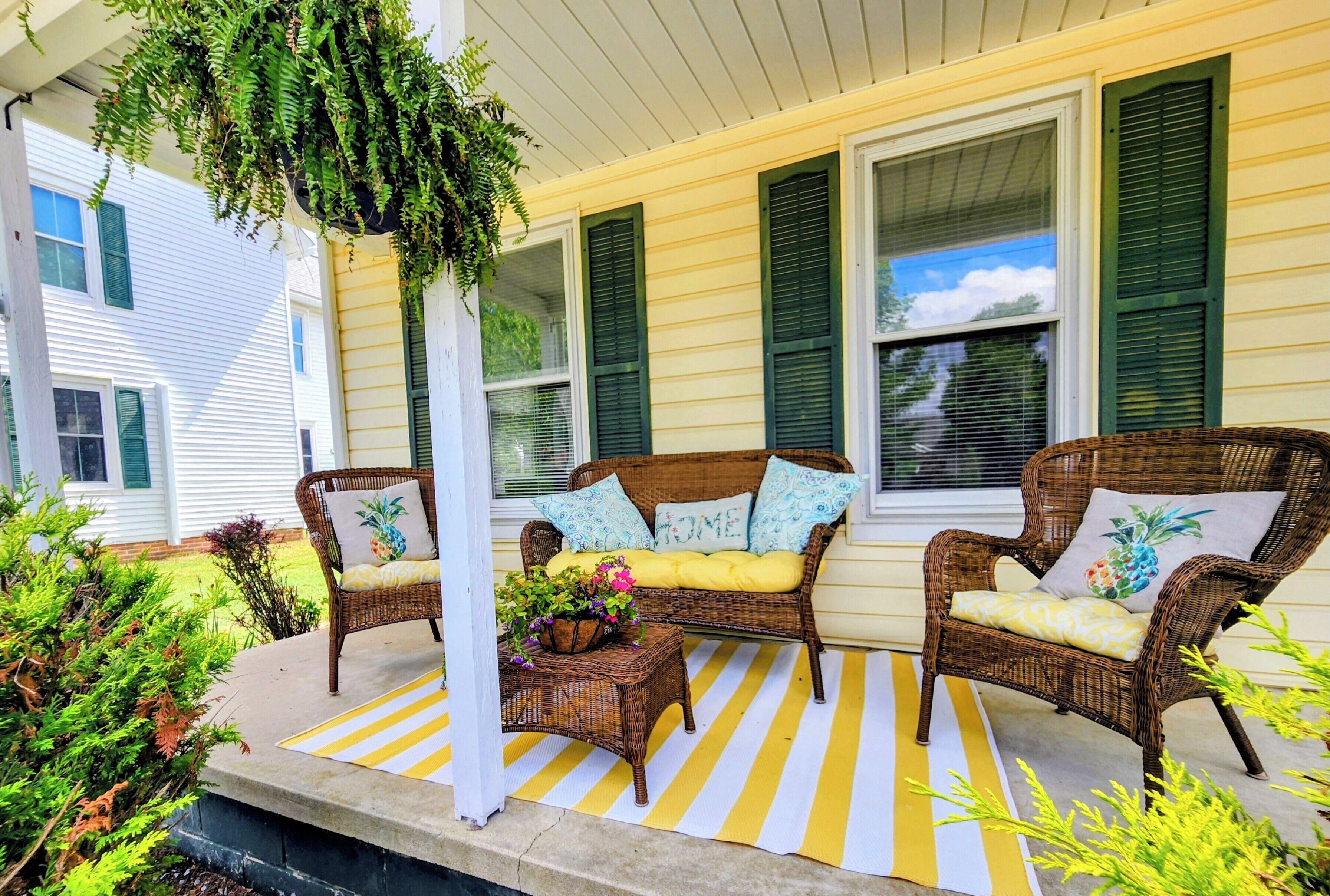 A porch with wicker furniture, a striped mat, and green foliage.