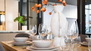 Mesa de comedor dispuesta con platos, tazones y copas de vino blancos, un jarrón blanco con flores naranjas y un interior moderno con una planta al fondo.