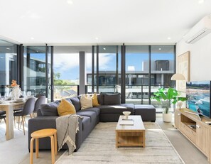 A modern living room with a sectional sofa, a coffee table, and a television. - KozyGuru | Parramatta | Sleek Skyline Retreat (Parramatta)
