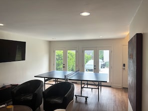 Ein Zimmer mit einer Tischtennisplatte, zwei Stühlen, einem Flachbildfernseher und einer zur Außenseite führenden Tür.