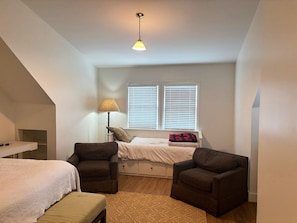 Ein Schlafzimmer mit einem Bett, zwei Sesseln, einer Lampe, einem Fenster mit Jalousien und einer Deckenleuchte.