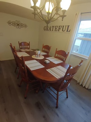 Ein Esszimmer mit Holztisch und -stühlen, einer Kristallleuchtre und einem Wandschild mit der Aufschrift 'GRATEFUL'.