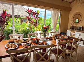 Un comedor con una mesa dispuesta para una comida, rodeada de sillas, un florero con flores y vistas a exuberante vegetación.