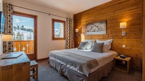 A bedroom with a large bed, wooden walls, a painting, a desk with a lamp, and a view of a snowy landscape. - Hotel Carlina Belle Plagne (La Plagne-Tarentaise)