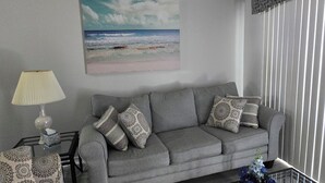 Um sofá cinzento com almofadas estampadas, uma mesa de centro de vidro com um estojo de lenços, uma luminária e um quadro de uma praia na parede.