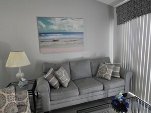 A grey sofa with patterned cushions, a glass coffee table with a tissue box, a lamp, and a framed beach painting on the wall. - Delightful 2-bedroom condo with AC in Myrtle Beach (Myrtle Beach)