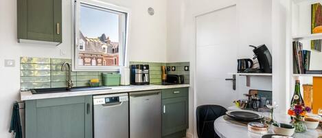Cucina compatta con finestra, mobili verdi, lavastoviglie e zona pranzo con tavolo apparecchiato per due.