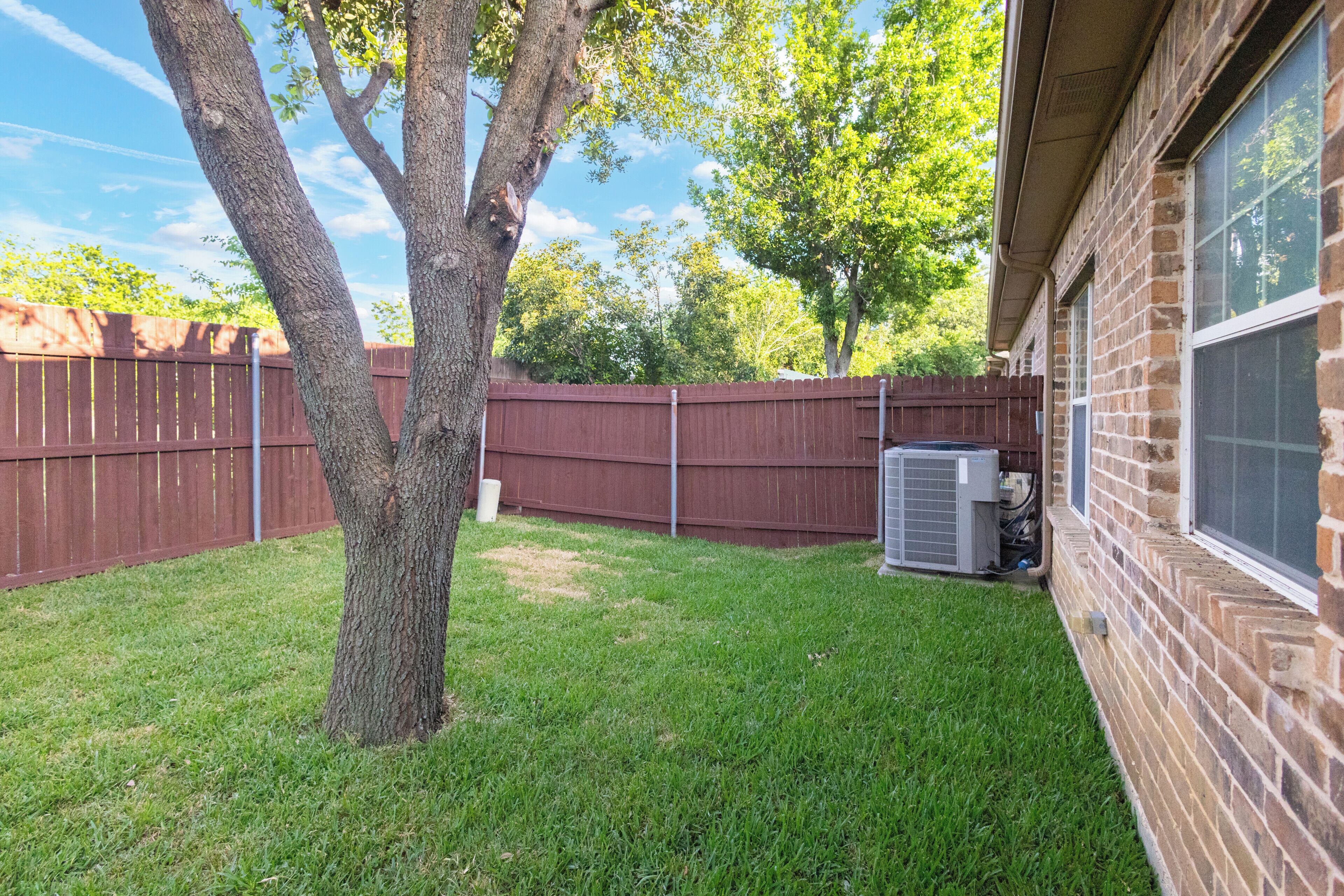 A backyard with a tree, a fence, and an air conditioning unit.